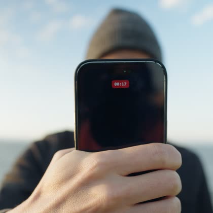 A phone held up screen-out, hiding the vlogger’s face behind a dark screen with a red recording timer.
